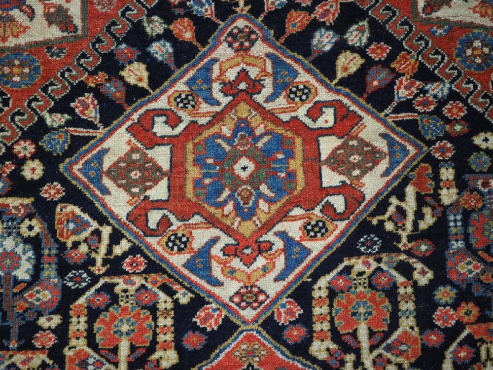 A hand-knotted wool rug featuring three diamond medallions on a dark navy field surrounded by geometric floral borders in red, ivory, and blue. View X