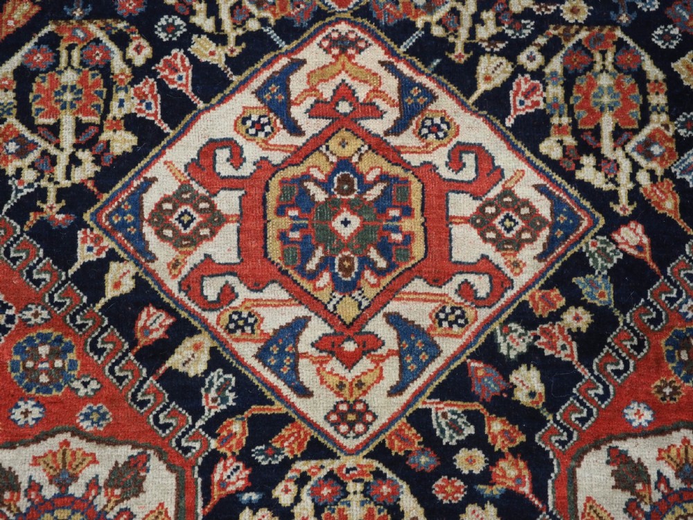 A hand-knotted wool rug featuring three diamond medallions on a dark navy field surrounded by geometric floral borders in red, ivory, and blue. View XI