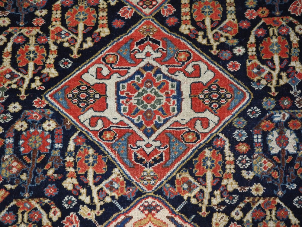 A hand-knotted wool rug featuring three diamond medallions on a dark navy field surrounded by geometric floral borders in red, ivory, and blue. View XIII