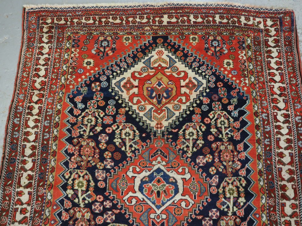 An antique hand-knotted Qashqai wool rug featuring a navy blue field with stacked diamond medallions surrounded by terracotta red borders and tribal geometric motifs. View III
