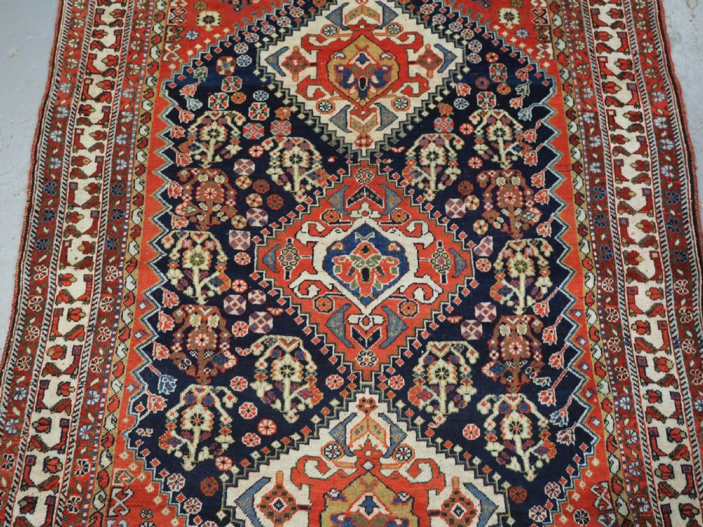An antique hand-knotted Qashqai wool rug featuring a navy blue field with stacked diamond medallions surrounded by terracotta red borders and tribal geometric motifs. View IV