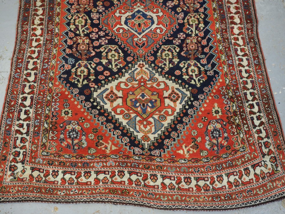 An antique hand-knotted Qashqai wool rug featuring a navy blue field with stacked diamond medallions surrounded by terracotta red borders and tribal geometric motifs. View V