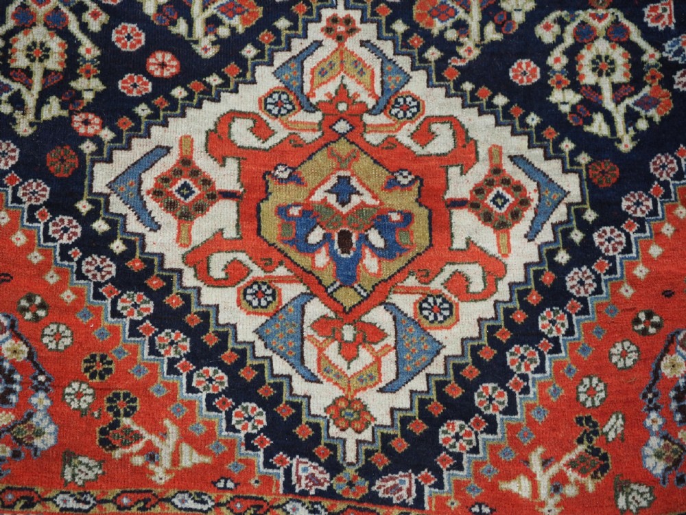 An antique hand-knotted Qashqai wool rug featuring a navy blue field with stacked diamond medallions surrounded by terracotta red borders and tribal geometric motifs. View IX