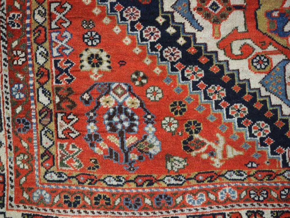 An antique hand-knotted Qashqai wool rug featuring a navy blue field with stacked diamond medallions surrounded by terracotta red borders and tribal geometric motifs. View X