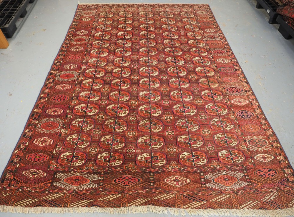 An antique Tekke Turkmen wool area rug featuring a repeating grid of octagonal gol motifs on a deep rust-red field with decorative geometric borders.