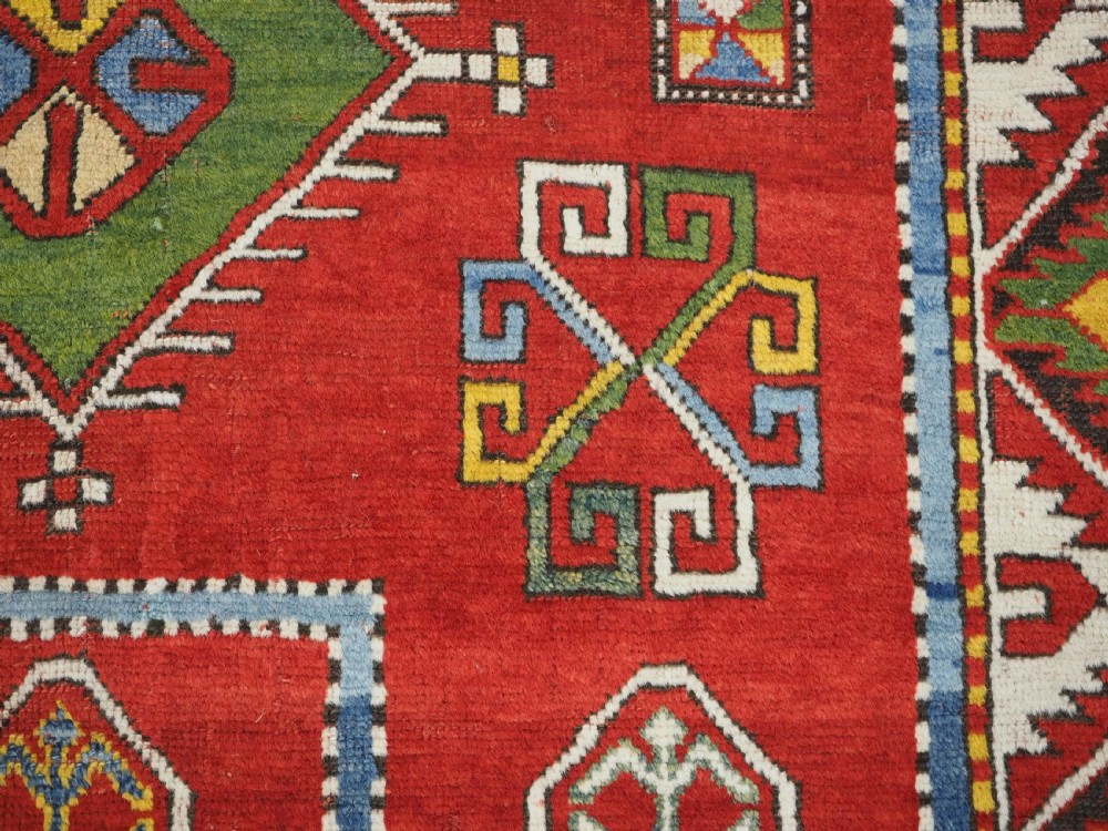 An antique hand-knotted Caucasian Fachralo Kazak prayer rug featuring a red field with green diamond medallions and a geometric border. View IX