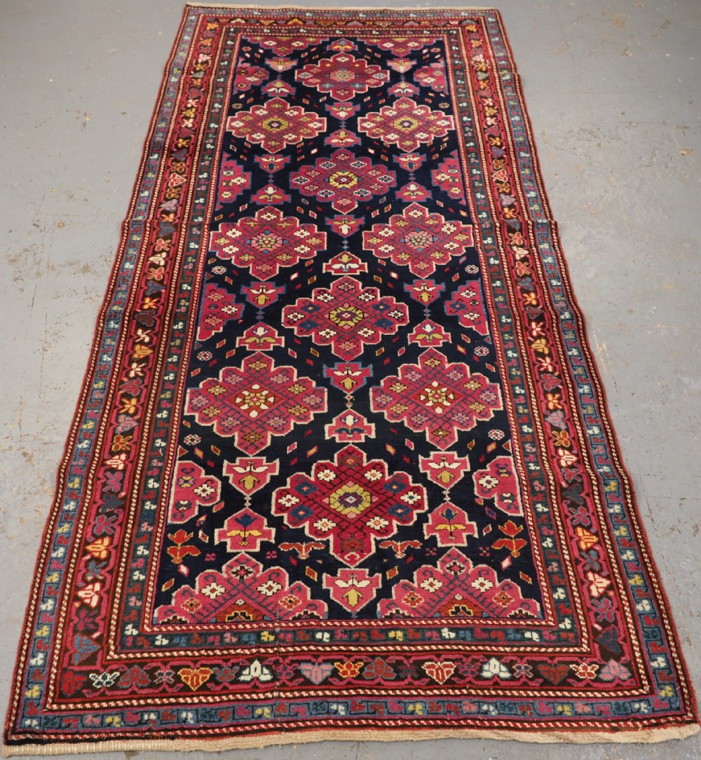 An antique long Caucasian runner rug featuring repeating pink and yellow geometric medallions on a dark navy field surrounded by a multi-band decorative border.