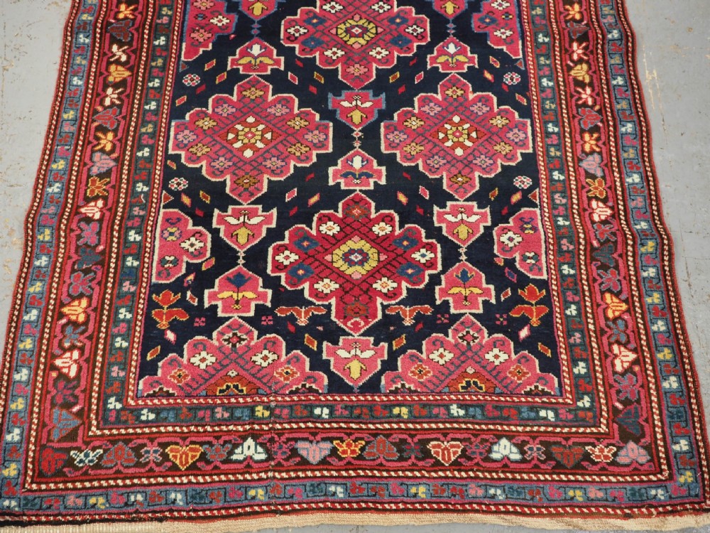 An antique long Caucasian runner rug featuring repeating pink and yellow geometric medallions on a dark navy field surrounded by a multi-band decorative border. View VI