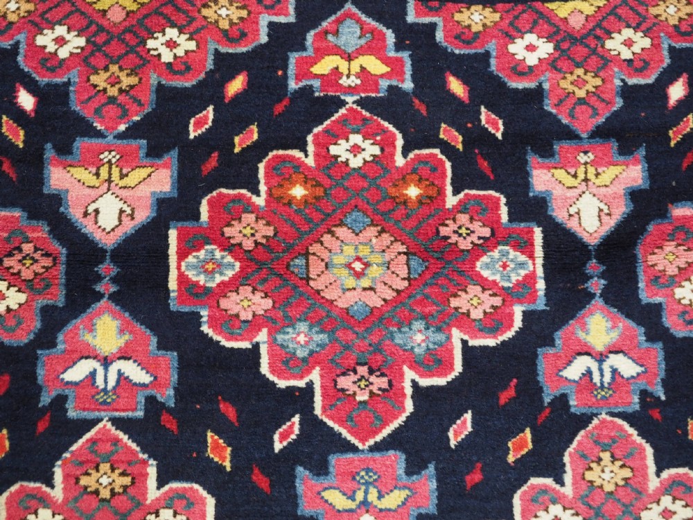 An antique long Caucasian runner rug featuring repeating pink and yellow geometric medallions on a dark navy field surrounded by a multi-band decorative border. View IX