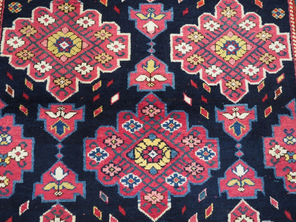An antique long Caucasian runner rug featuring repeating pink and yellow geometric medallions on a dark navy field surrounded by a multi-band decorative border. View X