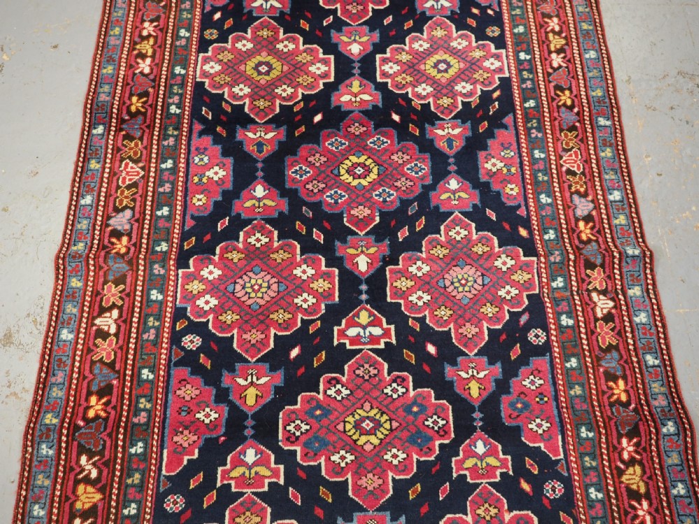 An antique long Caucasian runner rug featuring repeating pink and yellow geometric medallions on a dark navy field surrounded by a multi-band decorative border. View XVII