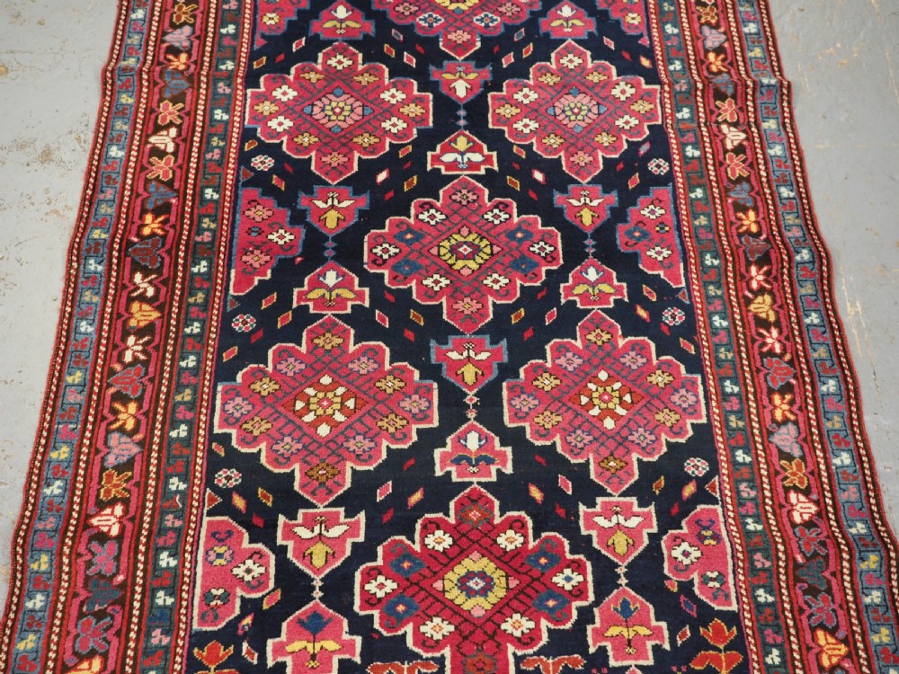 An antique long Caucasian runner rug featuring repeating pink and yellow geometric medallions on a dark navy field surrounded by a multi-band decorative border. View XVIII