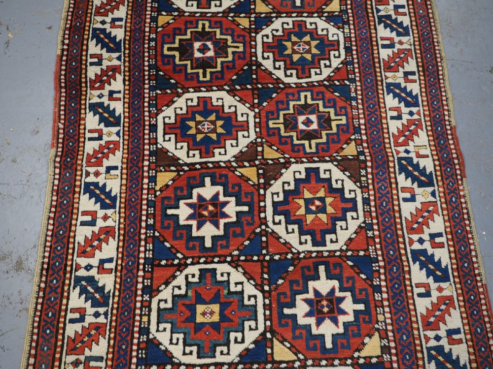 An antique Caucasian Moghan Kazak long wool rug featuring two vertical columns of octagonal geometric medallions in red, navy blue, cream, yellow, and green within a decorative patterned border. View IV