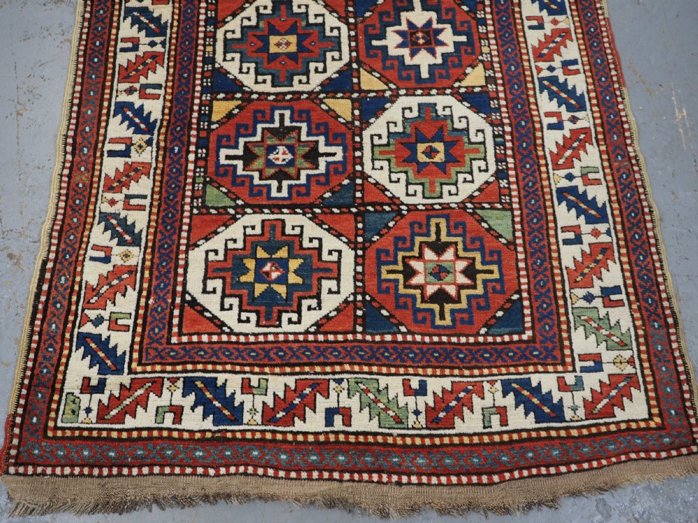 An antique Caucasian Moghan Kazak long wool rug featuring two vertical columns of octagonal geometric medallions in red, navy blue, cream, yellow, and green within a decorative patterned border. View V
