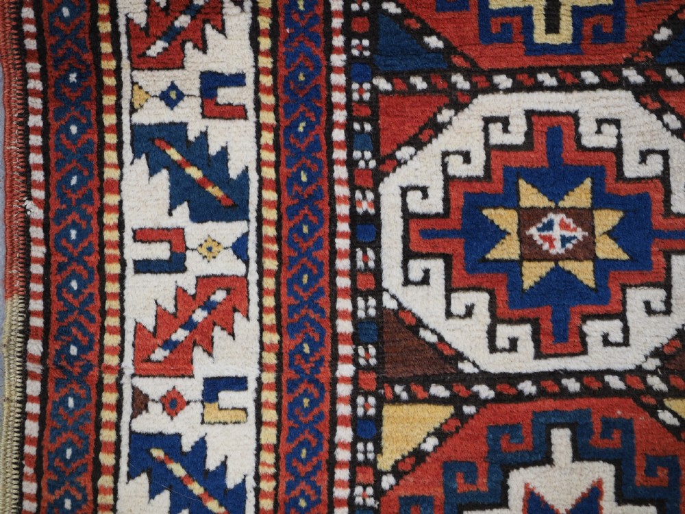 An antique Caucasian Moghan Kazak long wool rug featuring two vertical columns of octagonal geometric medallions in red, navy blue, cream, yellow, and green within a decorative patterned border. View VII