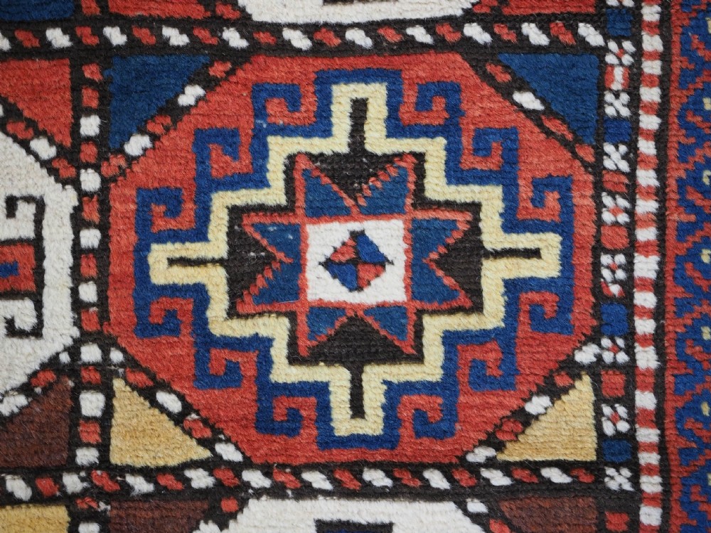 An antique Caucasian Moghan Kazak long wool rug featuring two vertical columns of octagonal geometric medallions in red, navy blue, cream, yellow, and green within a decorative patterned border. View IX