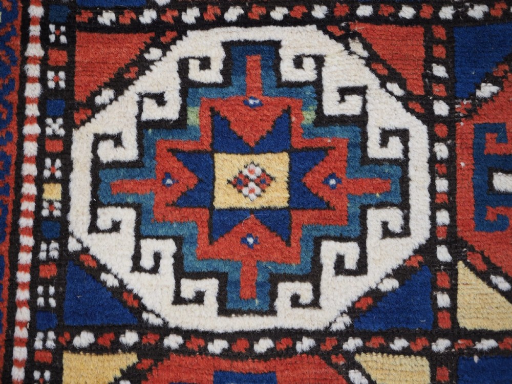 An antique Caucasian Moghan Kazak long wool rug featuring two vertical columns of octagonal geometric medallions in red, navy blue, cream, yellow, and green within a decorative patterned border. View X