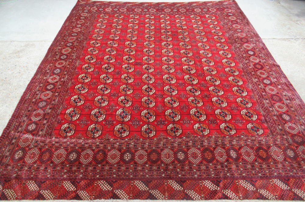 A large antique hand-knotted wool rug featuring repeating geometric gul motifs on a red field surrounded by multiple patterned borders.