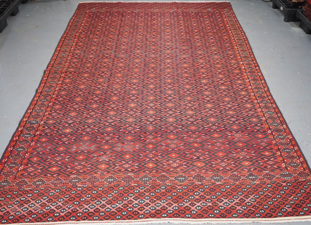 A large rectangular deep red wool flatweave carpet featuring an all-over diamond lattice pattern with detailed geometric borders and palas ends.