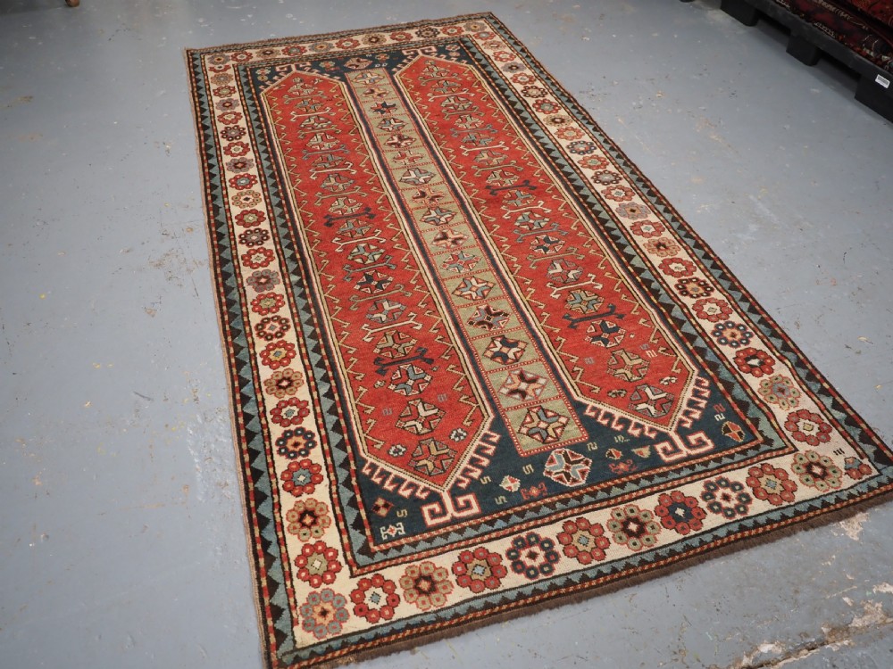 An elongated hand-knotted wool carpet featuring two central red field columns filled with geometric latch hook medallions framed by a multi-tiered cream and blue geometric border. View II