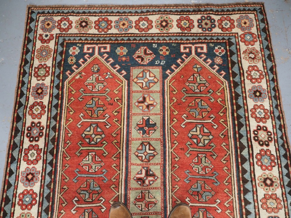 An elongated hand-knotted wool carpet featuring two central red field columns filled with geometric latch hook medallions framed by a multi-tiered cream and blue geometric border. View III