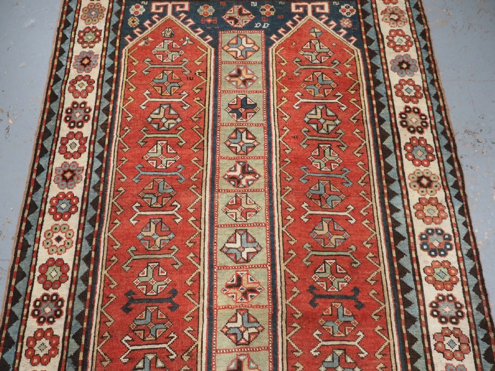 An elongated hand-knotted wool carpet featuring two central red field columns filled with geometric latch hook medallions framed by a multi-tiered cream and blue geometric border. View IV
