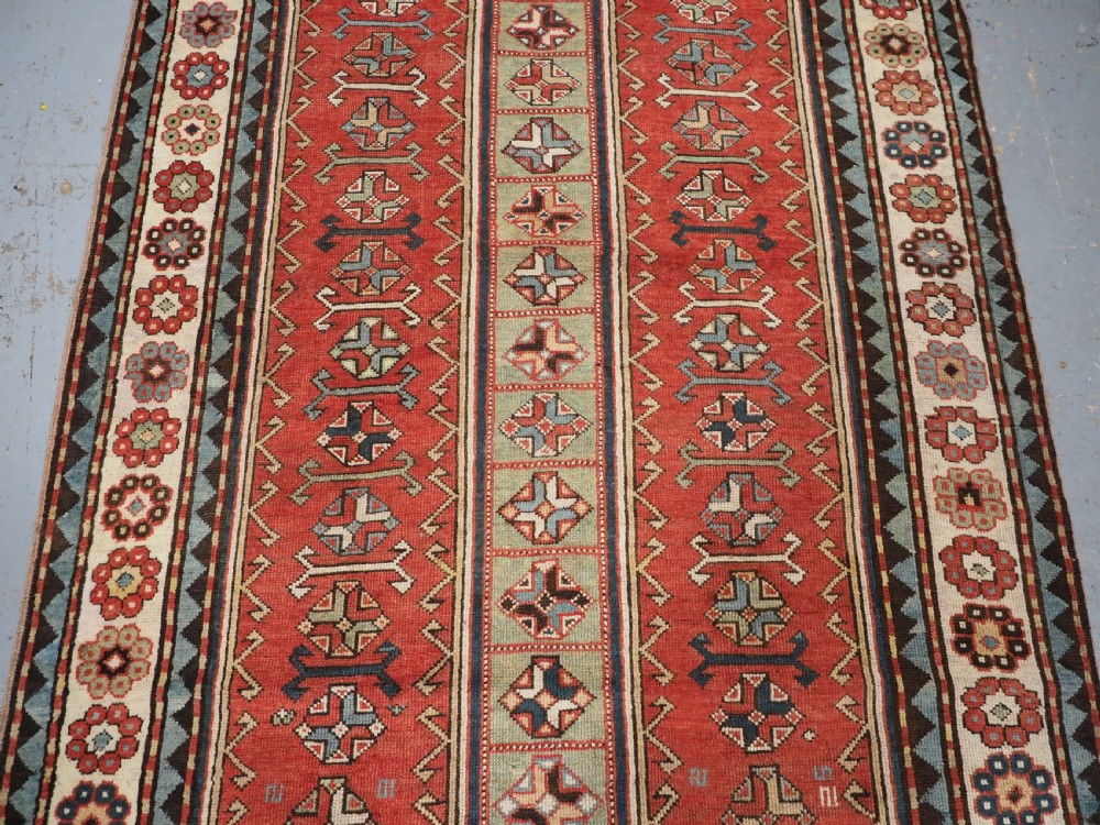 An elongated hand-knotted wool carpet featuring two central red field columns filled with geometric latch hook medallions framed by a multi-tiered cream and blue geometric border. View V