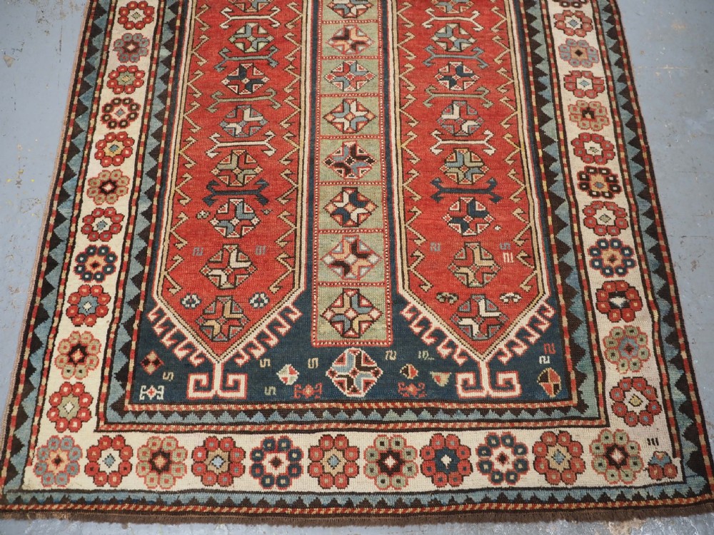 An elongated hand-knotted wool carpet featuring two central red field columns filled with geometric latch hook medallions framed by a multi-tiered cream and blue geometric border. View VI