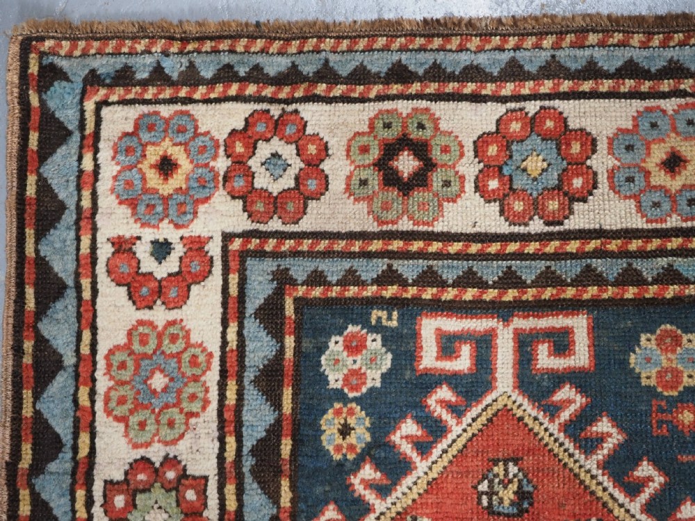 An elongated hand-knotted wool carpet featuring two central red field columns filled with geometric latch hook medallions framed by a multi-tiered cream and blue geometric border. View VII