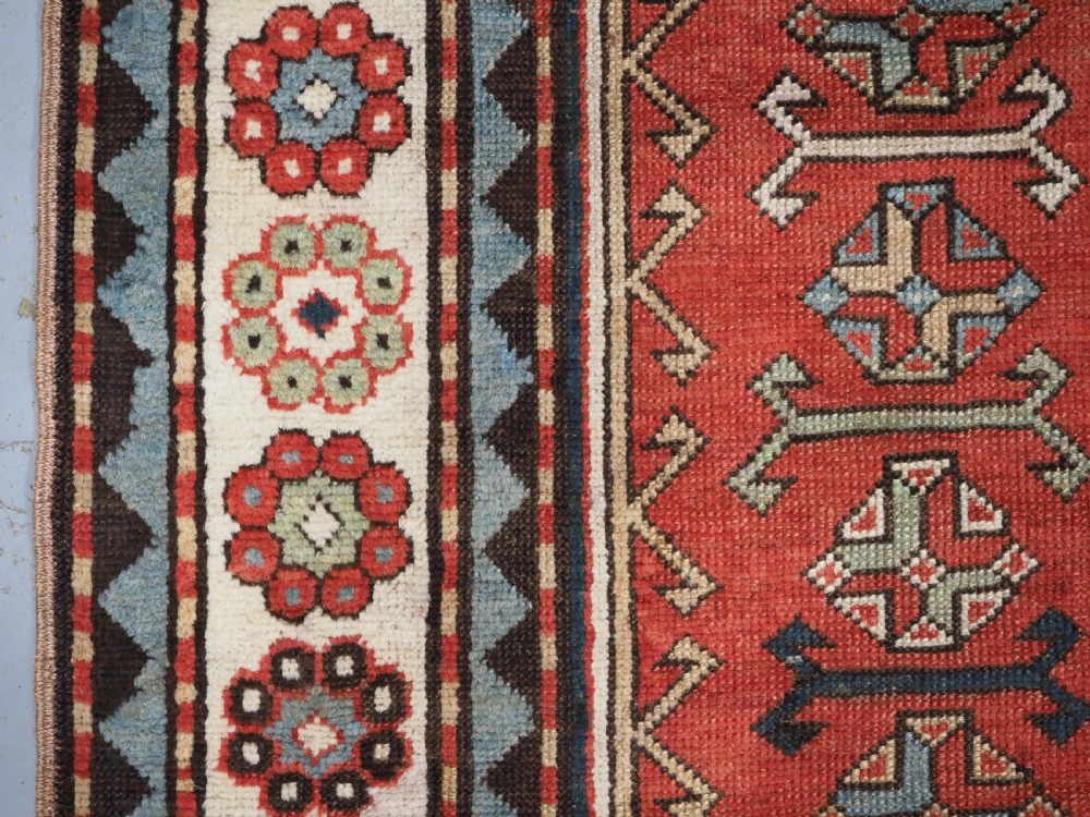 An elongated hand-knotted wool carpet featuring two central red field columns filled with geometric latch hook medallions framed by a multi-tiered cream and blue geometric border. View VIII