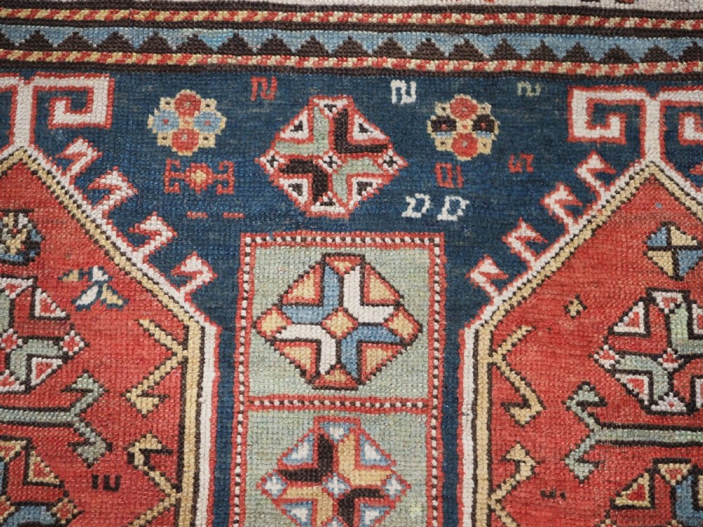 An elongated hand-knotted wool carpet featuring two central red field columns filled with geometric latch hook medallions framed by a multi-tiered cream and blue geometric border. View IX