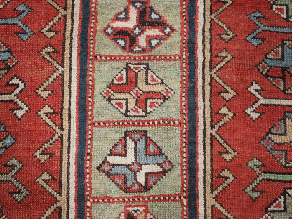 An elongated hand-knotted wool carpet featuring two central red field columns filled with geometric latch hook medallions framed by a multi-tiered cream and blue geometric border. View X