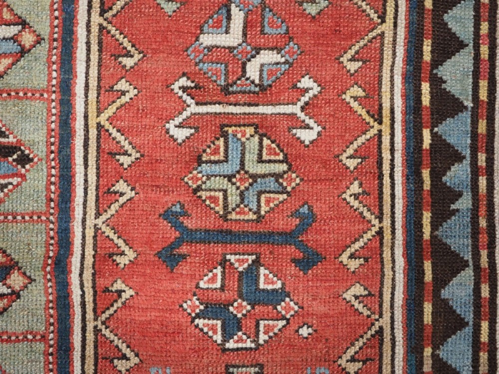 An elongated hand-knotted wool carpet featuring two central red field columns filled with geometric latch hook medallions framed by a multi-tiered cream and blue geometric border. View XI