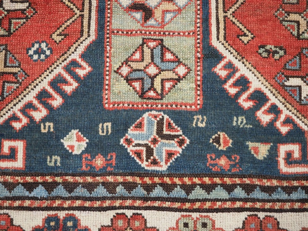 An elongated hand-knotted wool carpet featuring two central red field columns filled with geometric latch hook medallions framed by a multi-tiered cream and blue geometric border. View XII