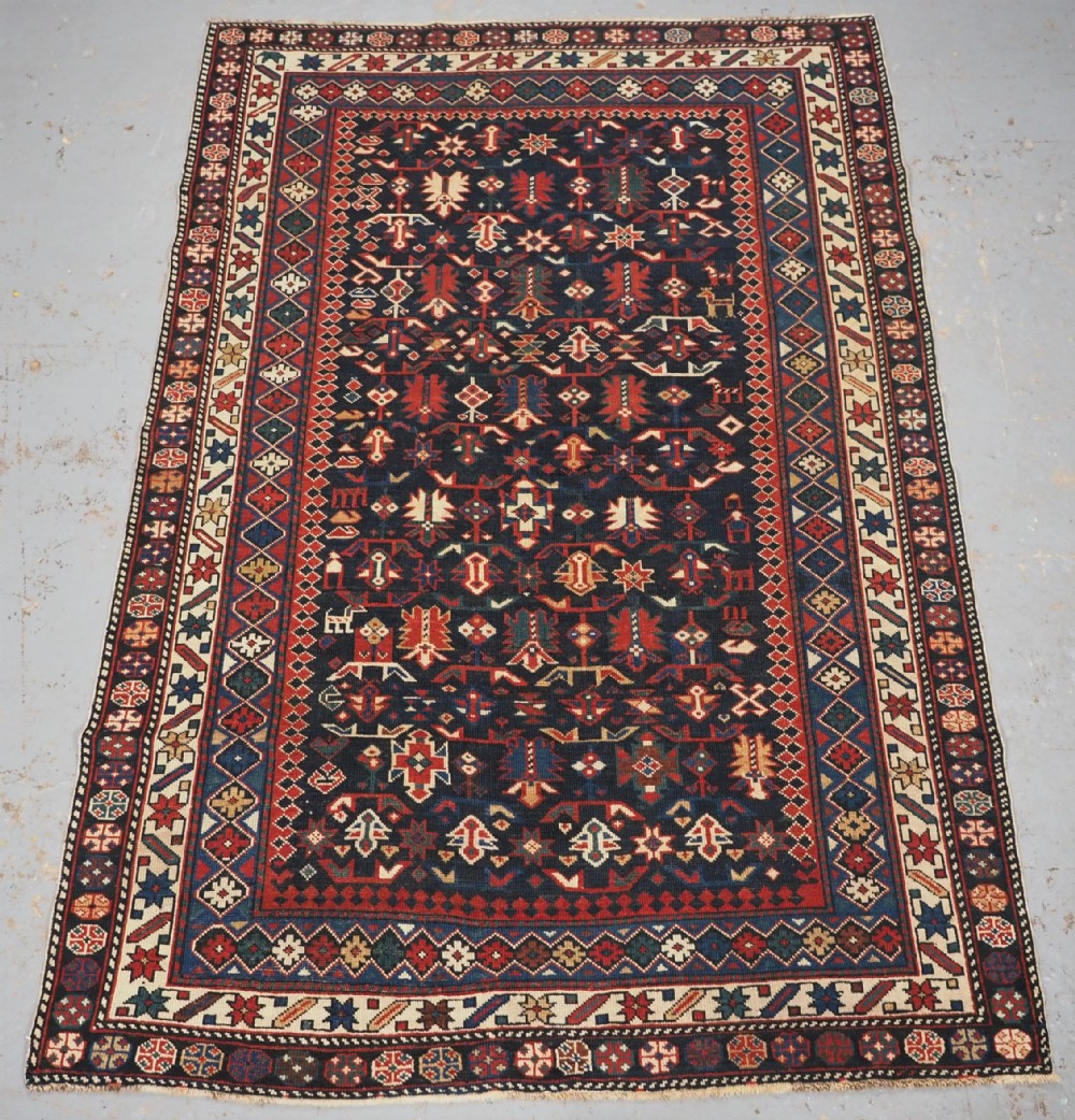 An antique hand-knotted wool rug featuring a dark navy central field filled with geometric tribal motifs, framed by multi-layered borders in red, cream, and blue hues.
