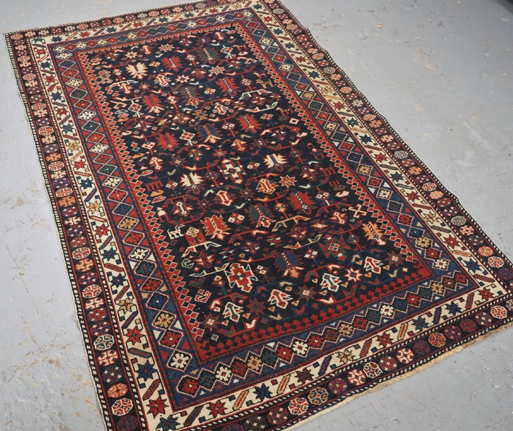 An antique hand-knotted wool rug featuring a dark navy central field filled with geometric tribal motifs, framed by multi-layered borders in red, cream, and blue hues. View II