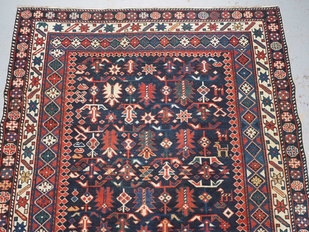 An antique hand-knotted wool rug featuring a dark navy central field filled with geometric tribal motifs, framed by multi-layered borders in red, cream, and blue hues. View III