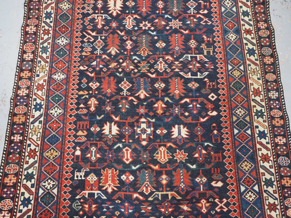 An antique hand-knotted wool rug featuring a dark navy central field filled with geometric tribal motifs, framed by multi-layered borders in red, cream, and blue hues. View IV