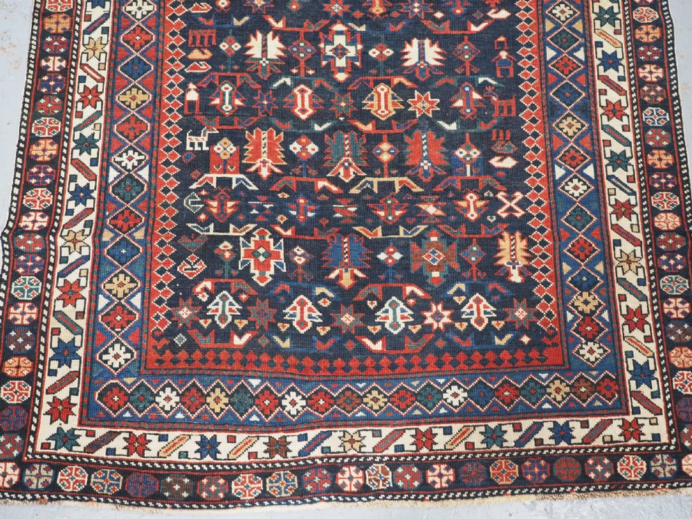 An antique hand-knotted wool rug featuring a dark navy central field filled with geometric tribal motifs, framed by multi-layered borders in red, cream, and blue hues. View V