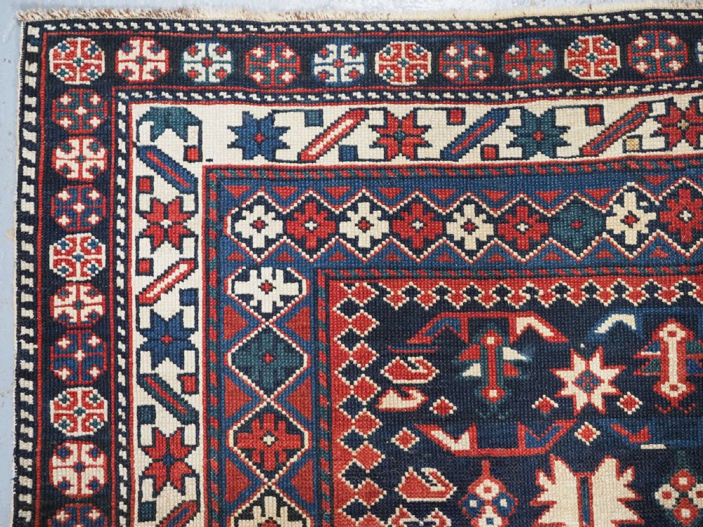 An antique hand-knotted wool rug featuring a dark navy central field filled with geometric tribal motifs, framed by multi-layered borders in red, cream, and blue hues. View VI