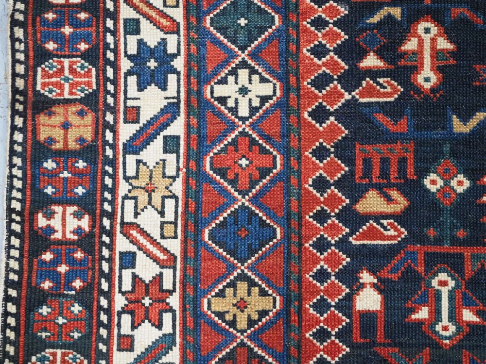 An antique hand-knotted wool rug featuring a dark navy central field filled with geometric tribal motifs, framed by multi-layered borders in red, cream, and blue hues. View VII