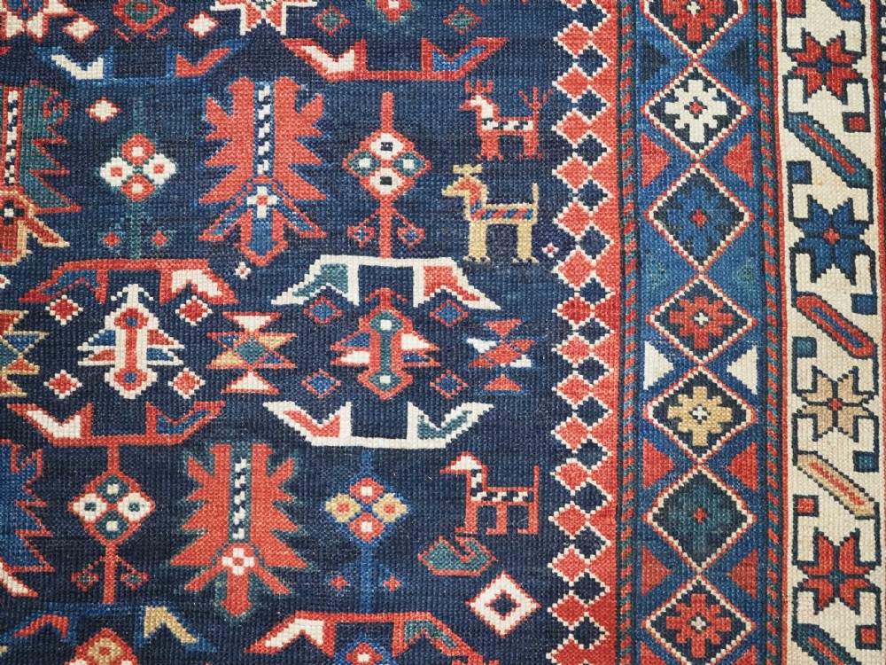 An antique hand-knotted wool rug featuring a dark navy central field filled with geometric tribal motifs, framed by multi-layered borders in red, cream, and blue hues. View IX