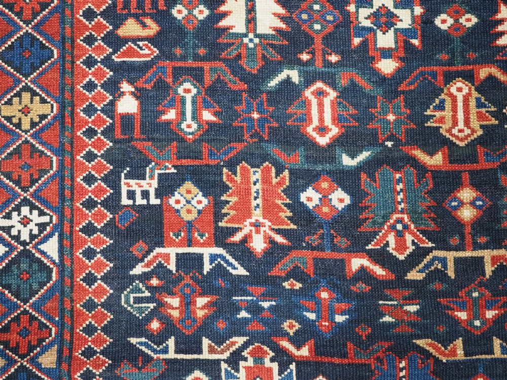 An antique hand-knotted wool rug featuring a dark navy central field filled with geometric tribal motifs, framed by multi-layered borders in red, cream, and blue hues. View X