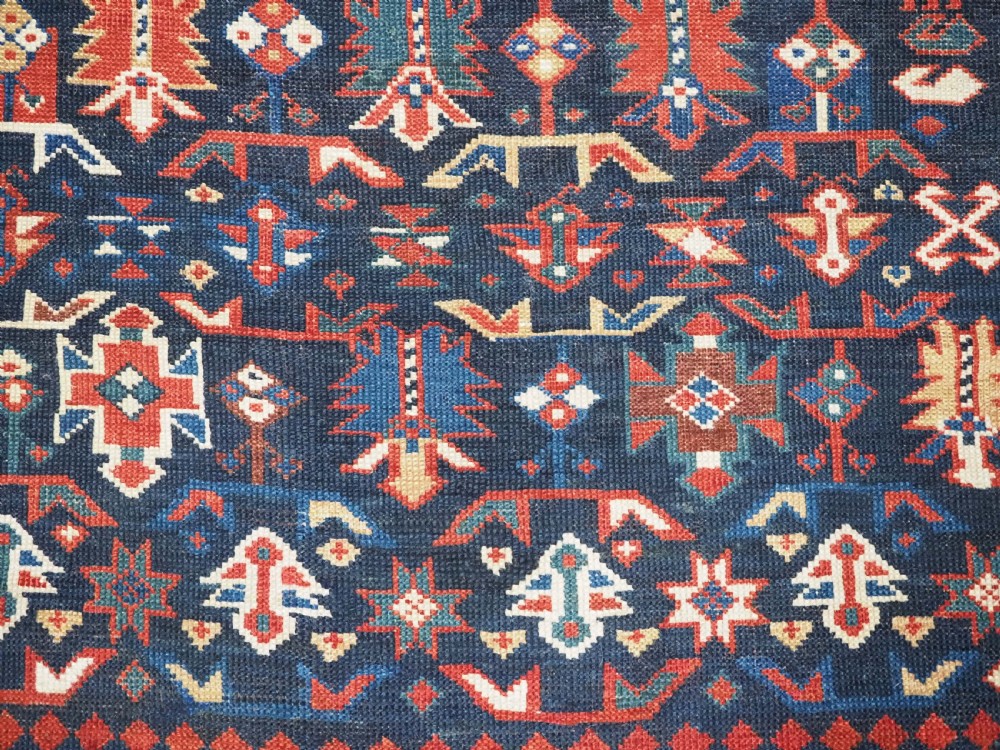 An antique hand-knotted wool rug featuring a dark navy central field filled with geometric tribal motifs, framed by multi-layered borders in red, cream, and blue hues. View XI