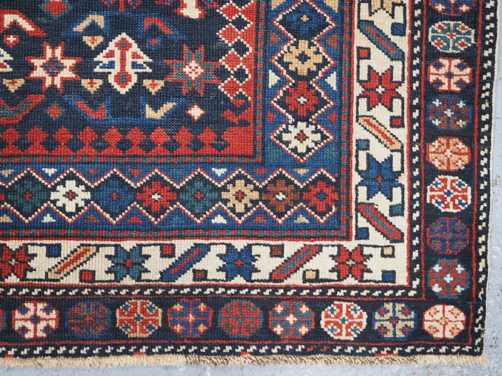 An antique hand-knotted wool rug featuring a dark navy central field filled with geometric tribal motifs, framed by multi-layered borders in red, cream, and blue hues. View XII