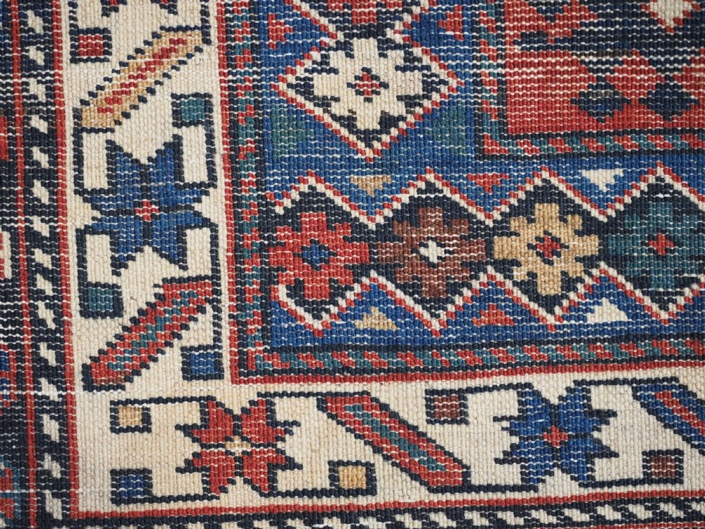 An antique hand-knotted wool rug featuring a dark navy central field filled with geometric tribal motifs, framed by multi-layered borders in red, cream, and blue hues. View XIII