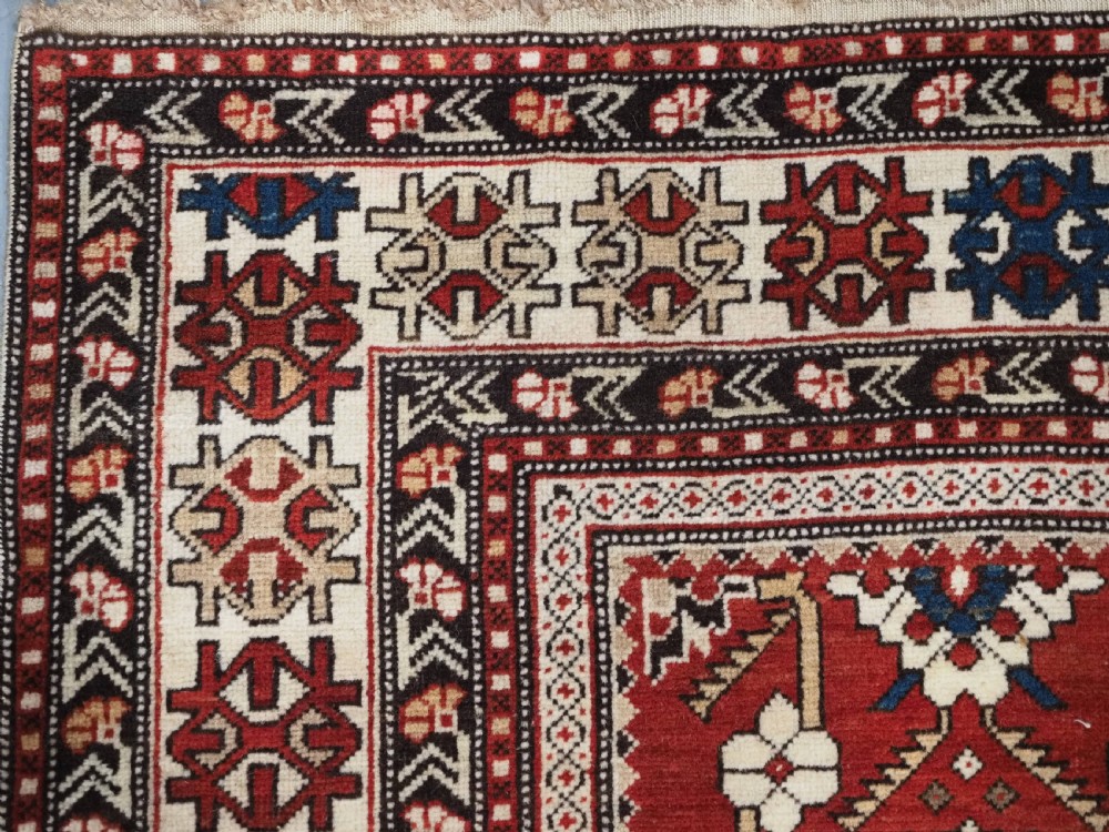 An antique hand-knotted Shirvan wool rug featuring a red field with an intricate cream floral lattice pattern framed by multi-layered geometric borders. View V