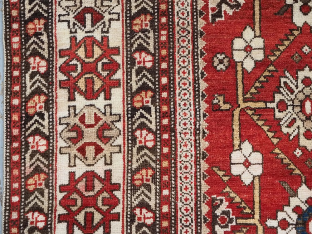 An antique hand-knotted Shirvan wool rug featuring a red field with an intricate cream floral lattice pattern framed by multi-layered geometric borders. View VI