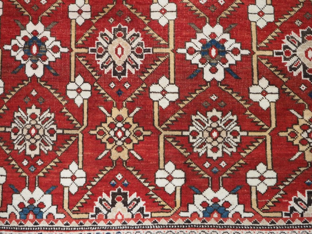 An antique hand-knotted Shirvan wool rug featuring a red field with an intricate cream floral lattice pattern framed by multi-layered geometric borders. View IX