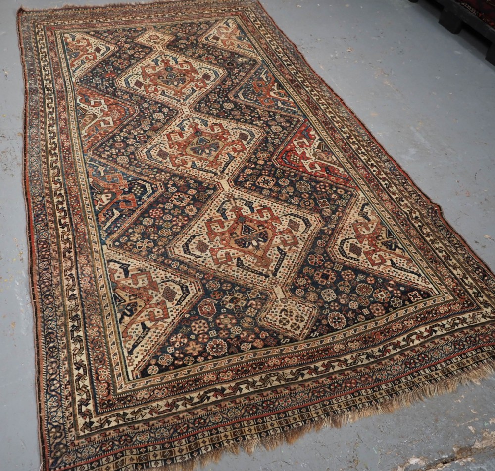 An antique rectangular wool rug featuring three diamond-shaped medallions along a central dark blue field surrounded by geometric borders in terracotta, cream, and blue tones. View II